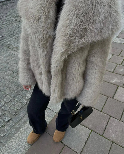 Grauer Kunstpelzmantel, schwarze Handtasche, braune Stiefel auf gepflastertem Gehweg.