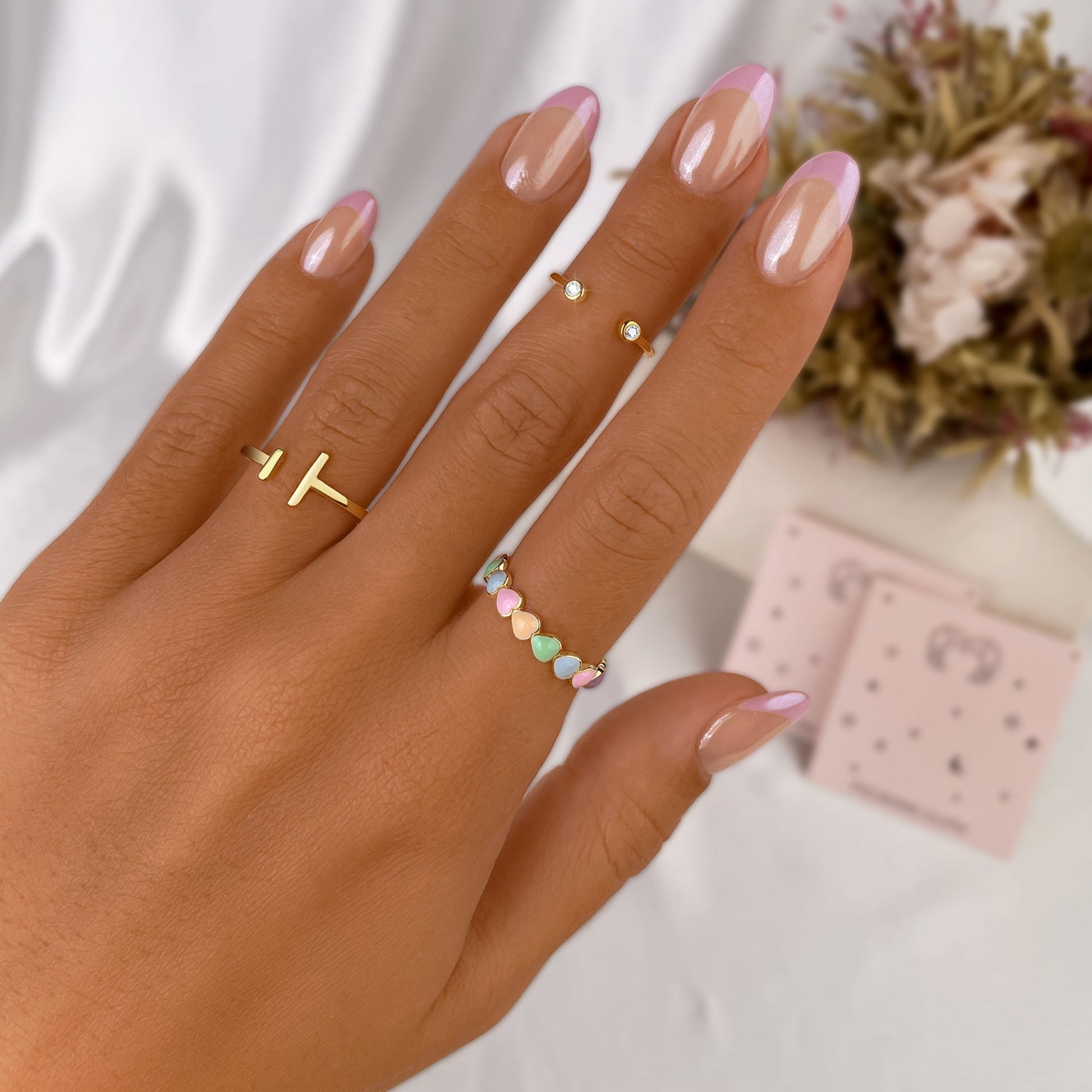 Hand mit goldenen Ringen und Herz-Design, rosa French Maniküre, Blumen im Hintergrund.