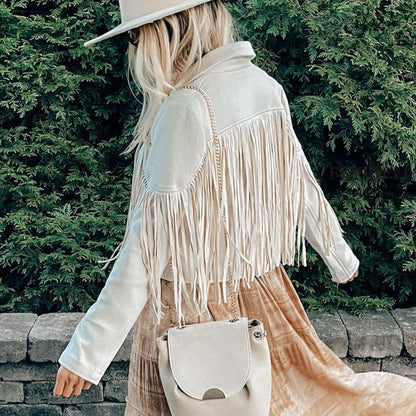 Frau in weißer Fransenjacke und Hut, beige Tasche, vor grüner Hecke.