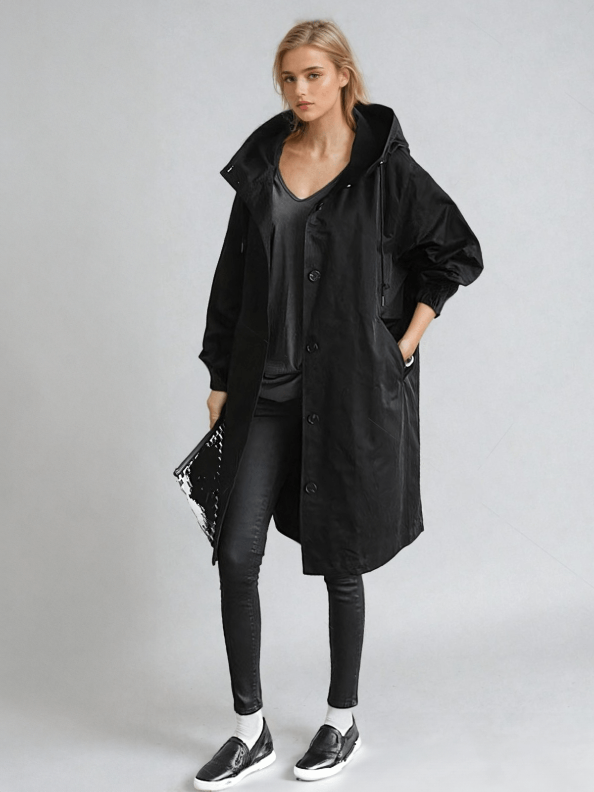Helfigur av kvinna i svart långkappa med huva, svart topp, svarta jeans, sneakers och blank clutch. 2