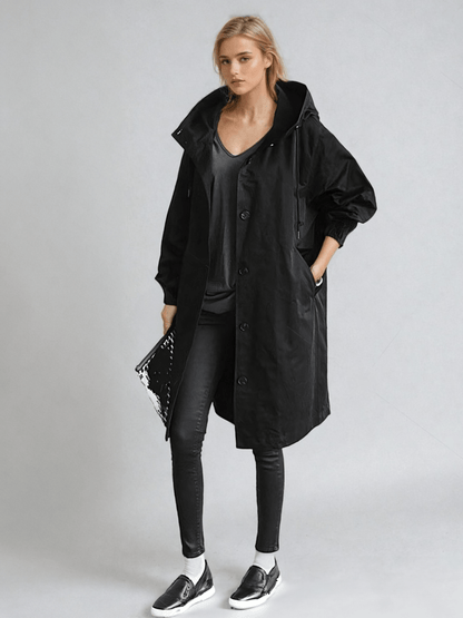 Helfigur av kvinna i svart långkappa med huva, svart topp, svarta jeans, sneakers och blank clutch. 2