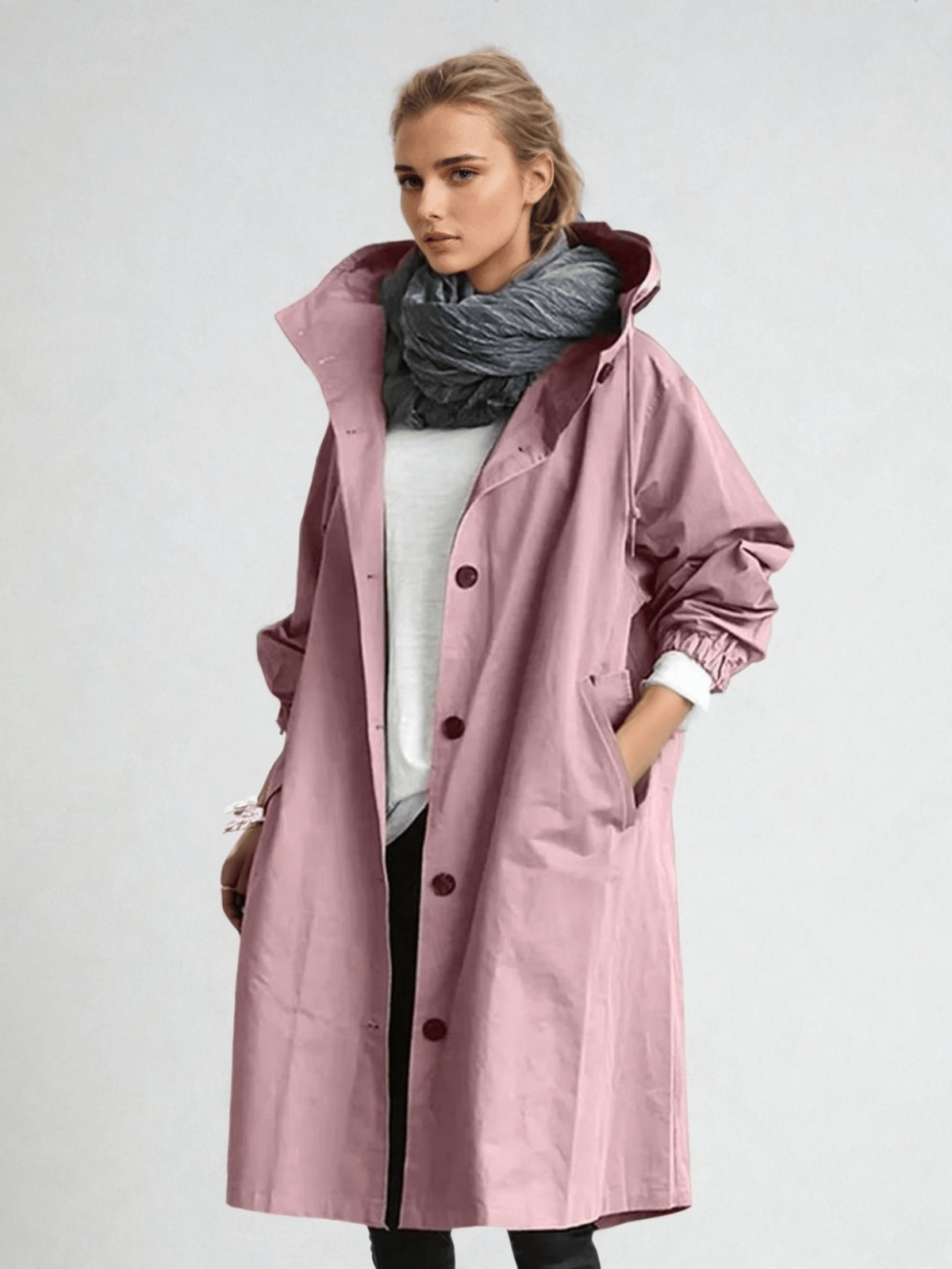 Kvinna i rosa knäppt kappa med huva, vit tröja och grå halsduk, ena handen i fickan. 6