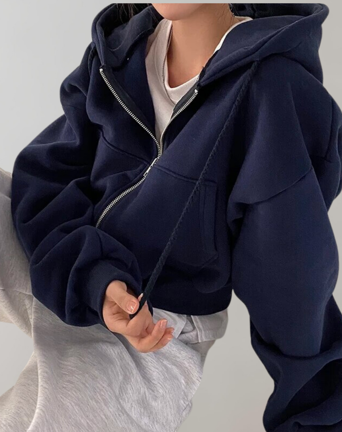 Närbild på marinblå hoodie med dragkedja och ficka, bärd över vit tröja och ljusgrå byxor, ena handen håller i dragsnöret. 3