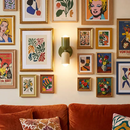 Tänd vägglampa i olivgrönt och gräddvitt mitt i ett tätt galleri av inramade posters ovanför en orange sammetssoffa. 6