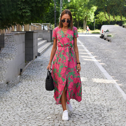Frau in rosa-blauem Blumenkleid auf gepflasterter Straße, trägt schwarze Tasche, weiße Sneaker.