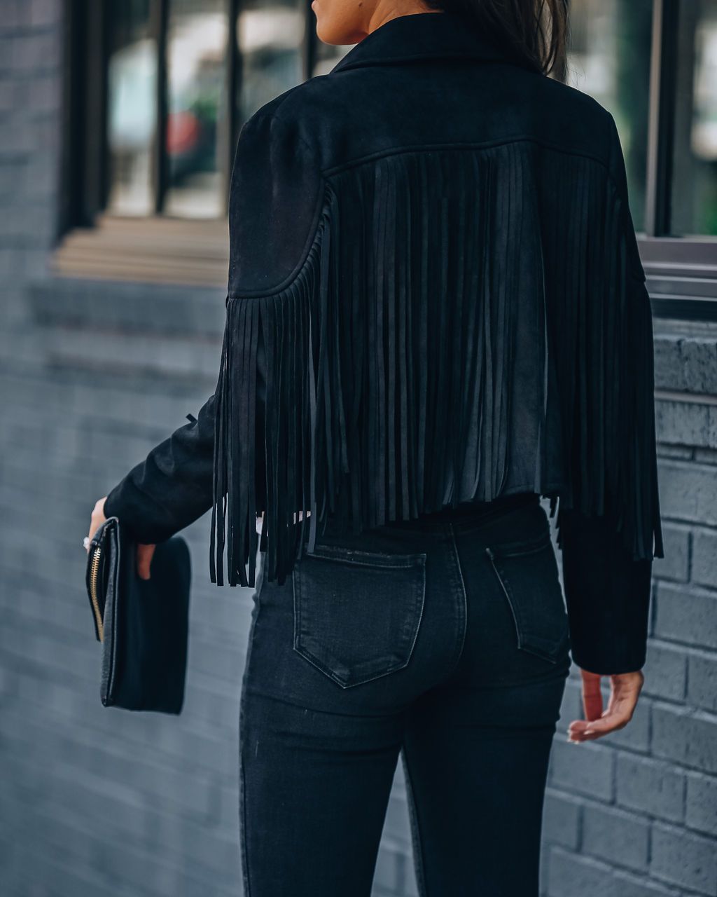 Frau in schwarzer Fransenjacke aus Wildleder, hält schwarze Clutch, modisch, urbaner Stil.