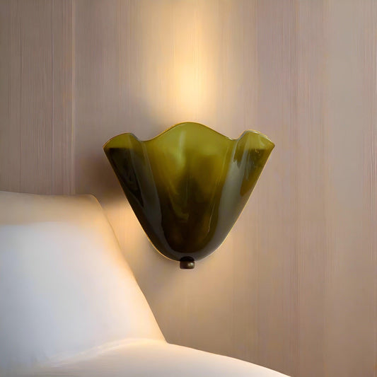 Närbild på tänd olivgrön glasvägglampa med vågig kant, monterad på en ljus panelvägg bredvid ett vitt kuddhörn. 2