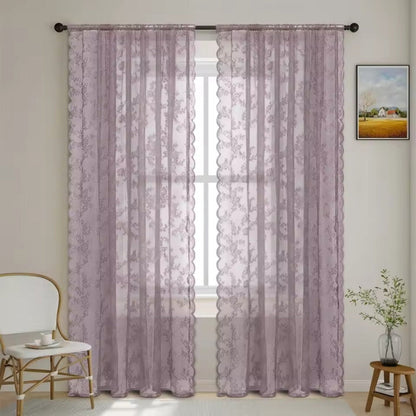 Dämpat mauve genomskinliga spetsgardiner med blommönster och vågig kant hänger vid ett fönster bredvid en vit stol och ett litet sidobord. 11
