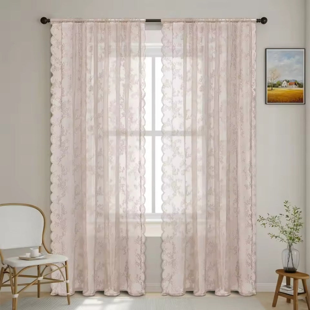 Ljust rosabeige genomskinliga spetsgardiner med fint blommönster och vågig kant hänger vid ett fönster intill en vit stol och ett litet bord med växt. 9