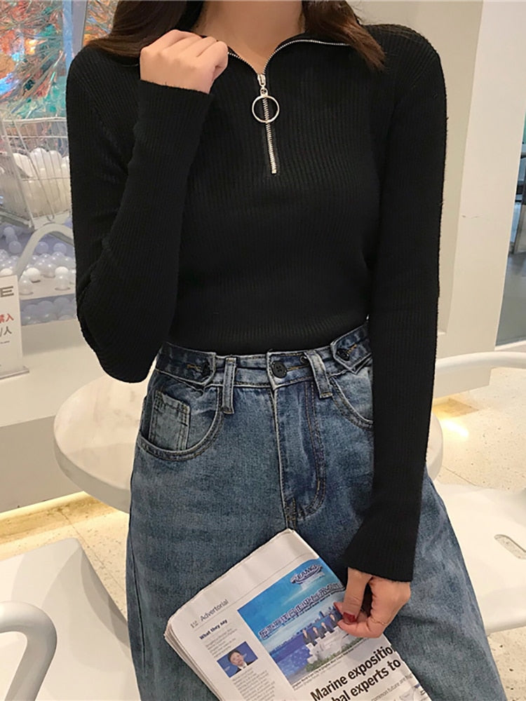 Frau in schwarzem Pullover mit Reißverschluss, blaue Jeans, hält Zeitung in modernem Raum.