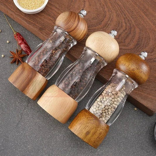 Drei Holz-Pfeffermühlen mit Glasgehäuse auf grauem Untergrund, gefüllt mit Pfefferkörnern.
