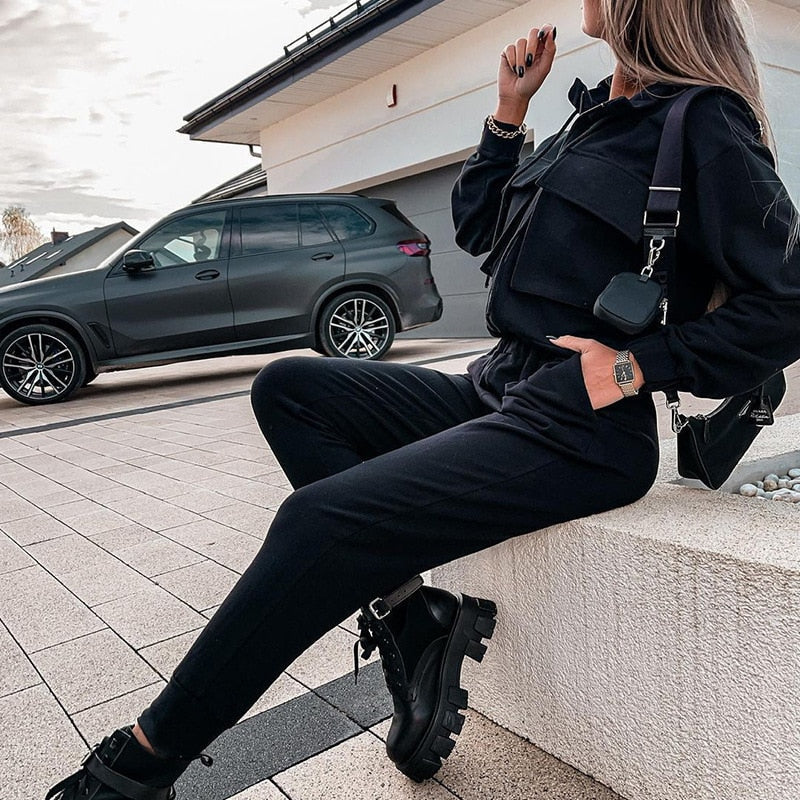 Frau in schwarzem Jogginganzug vor modernem SUV, modische Freizeitkleidung, urbane Umgebung.