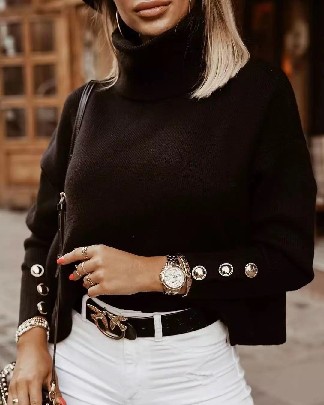 Frau in schwarzem Rollkragenpullover mit Knöpfen, weißer Hose, Uhr und Hut, modisch gekleidet.