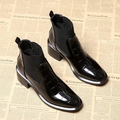 Schwarze Lackleder-Stiefeletten mit Krokodilprägung auf Zeitungshintergrund.