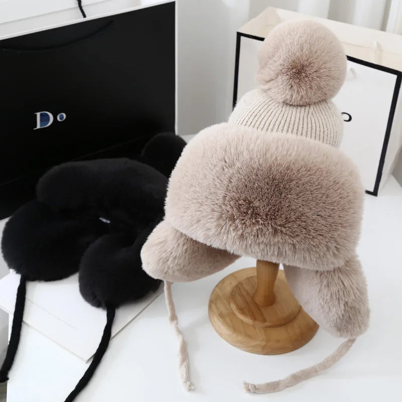 Beige Wintermütze mit Bommel, Kunstfell, warm, stilvoll, auf Holzständer, neben schwarzer Mütze.