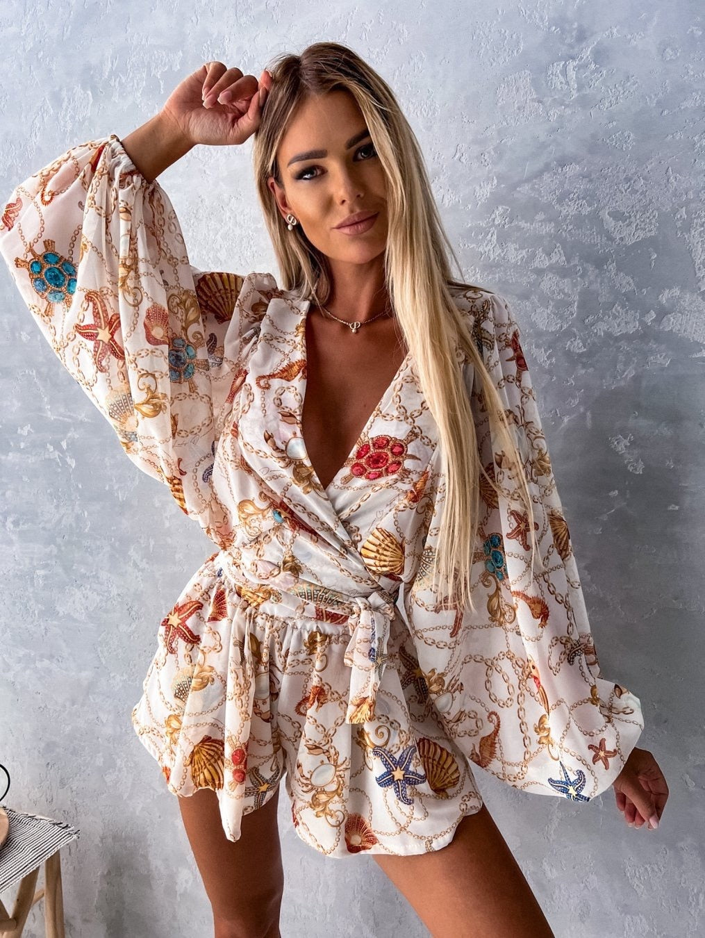 Frau in weißem, gemustertem Sommerkleid mit Muschel- und Kettenmotiv vor grauer Wand.