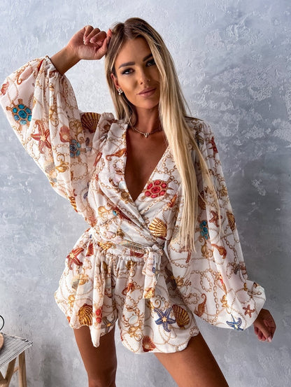 Frau in weißem, gemustertem Sommerkleid mit Muschel- und Kettenmotiv vor grauer Wand.