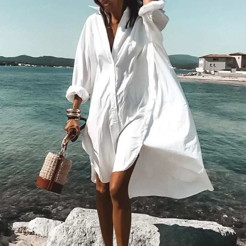 Frau in weißem Sommerkleid am Strand, trägt Korbtasche, Meer im Hintergrund.