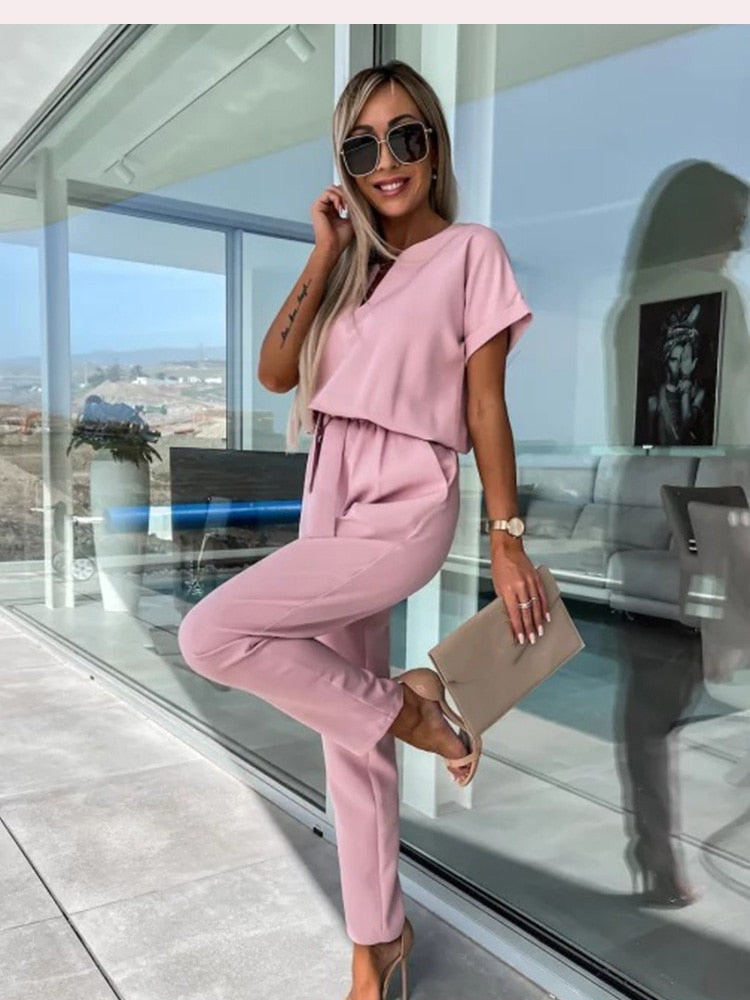 Frau in rosa Jumpsuit, Sonnenbrille, beige Clutch, moderne Terrasse, stilvolle Sommermode.