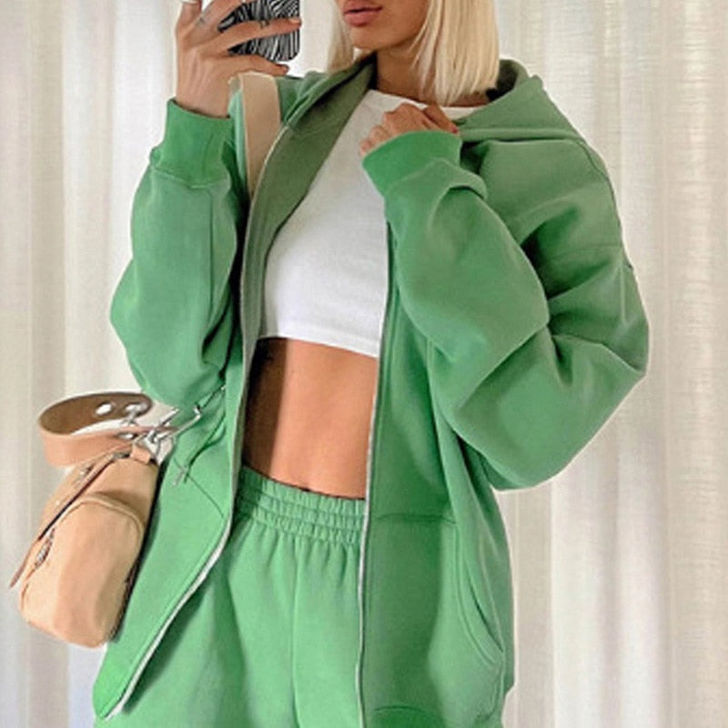 Frau in grünem Jogginganzug mit Kapuze, weißem Top, beige Tasche, Selfie im Spiegel.