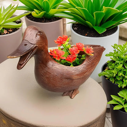 Entenförmiger Blumentopf aus Holz mit roten Blumen, umgeben von grünen Pflanzen in Töpfen.