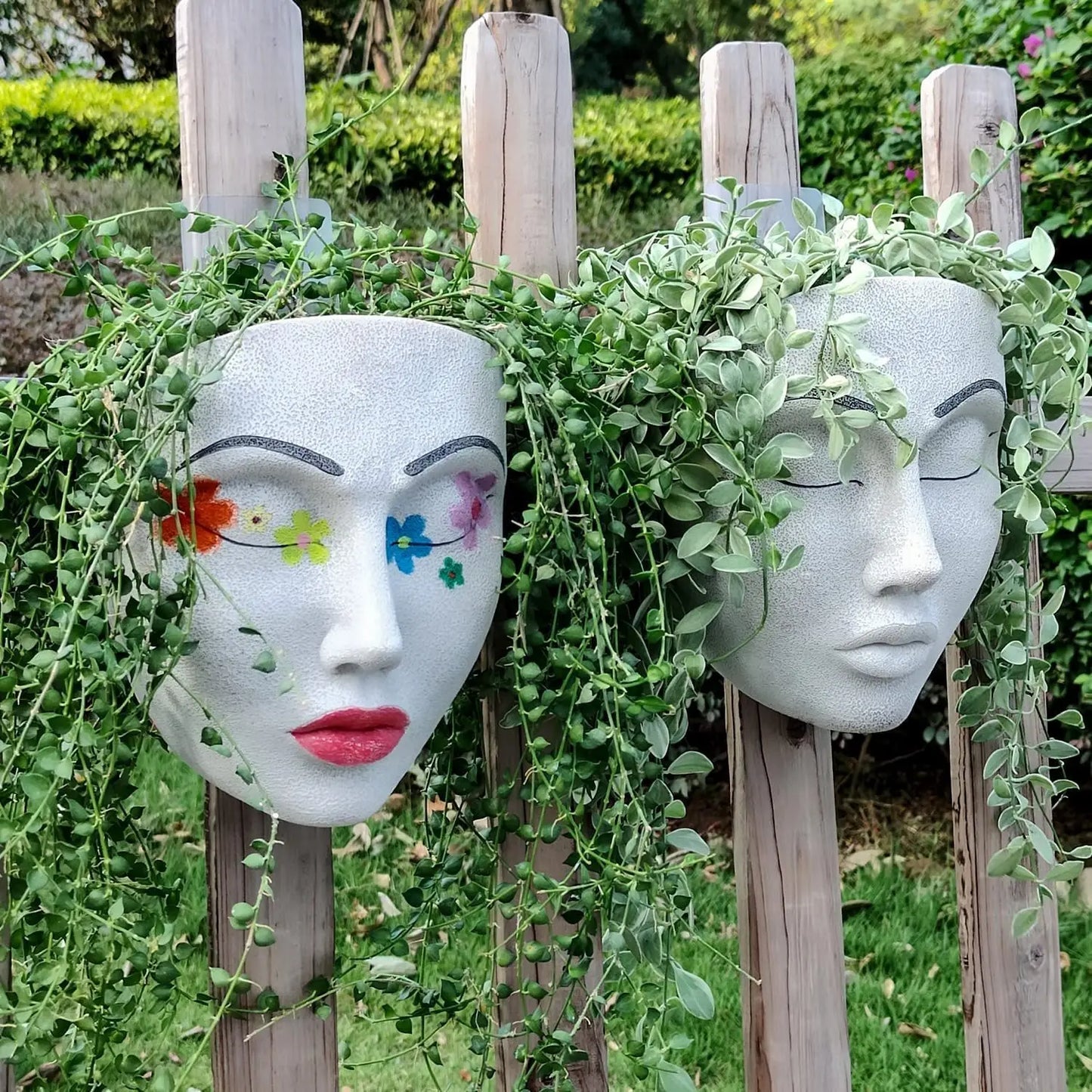 Zwei weiße Pflanzgefäße in Gesichtsform mit Blumenmotiven und hängenden Pflanzen auf Holzzaun.