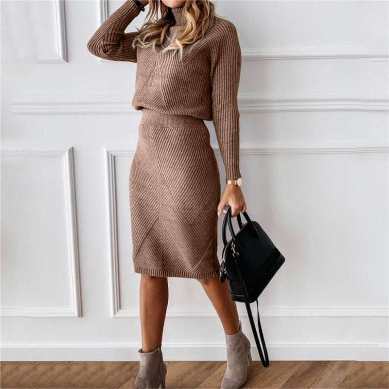 Frau in braunem Strickkleid mit Handtasche und Stiefeletten vor weißer Wand.