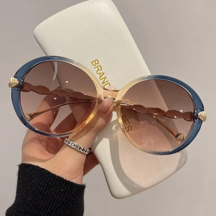 Große runde Sonnenbrille mit blau-goldenem Rahmen und braunen Gläsern, in Etui gehalten.