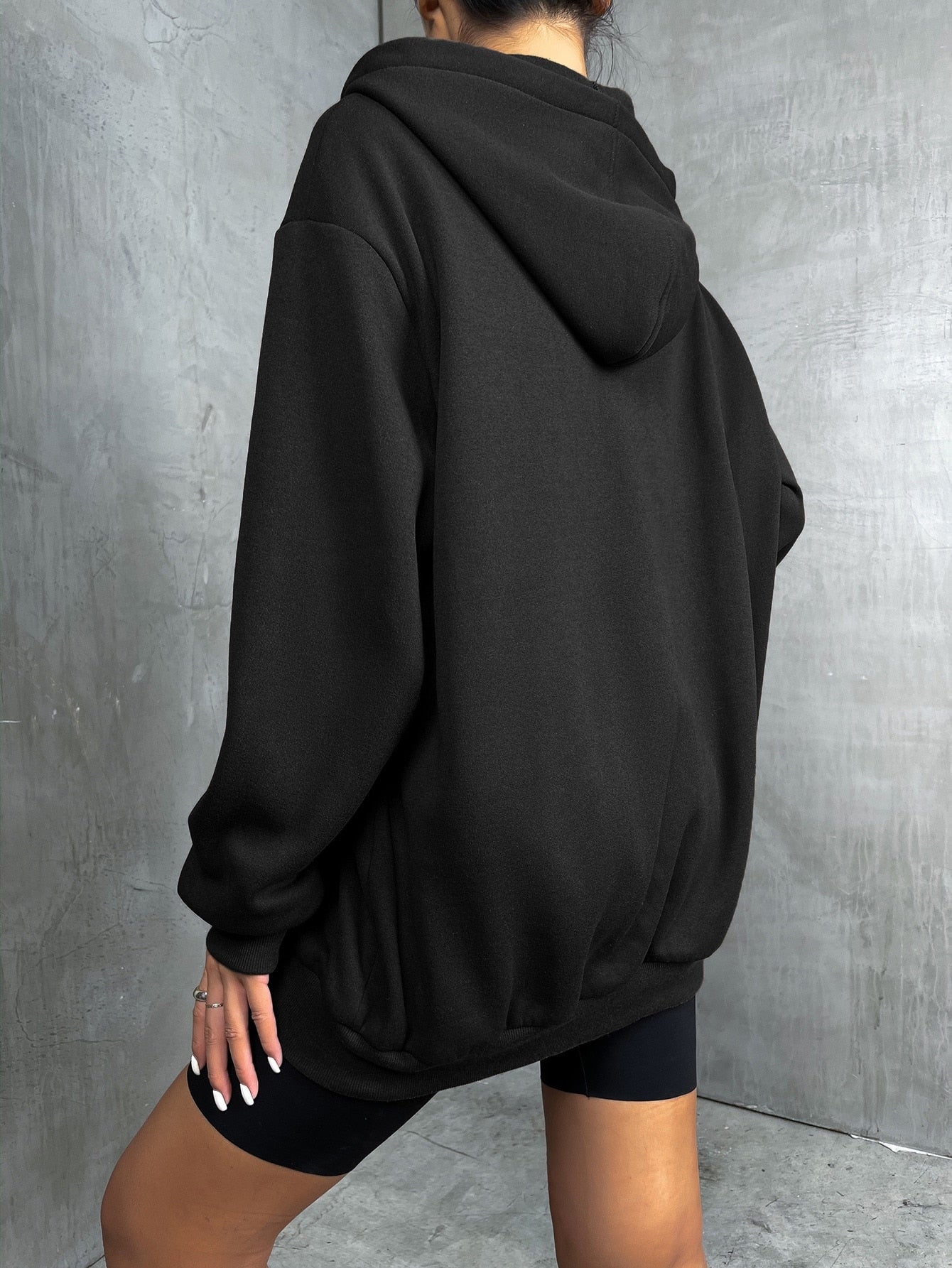 Frau in schwarzem Oversize-Hoodie und schwarzen Shorts vor grauer Wand.