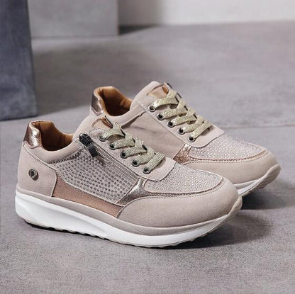 Ett par beige sneakers med guldfärgade glittersnören, glänsande detaljer och blixtlås, placerade på ett grått golv utan modell. 2