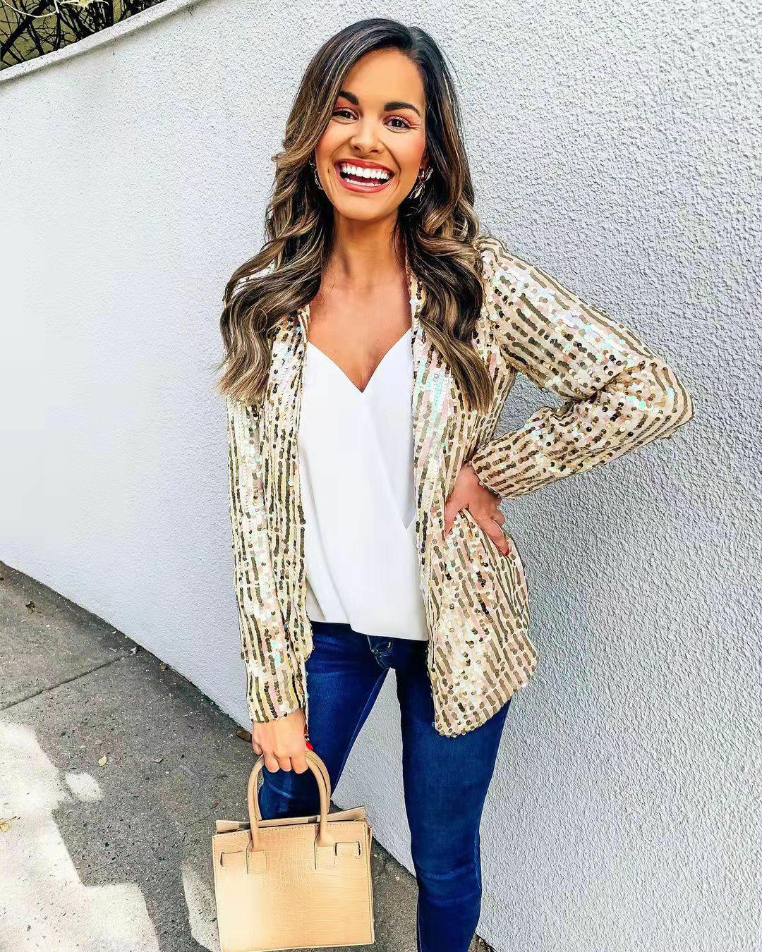 Frau in goldener Paillettenjacke, weißem Top, blauer Jeans, hält beige Handtasche, lächelt.
