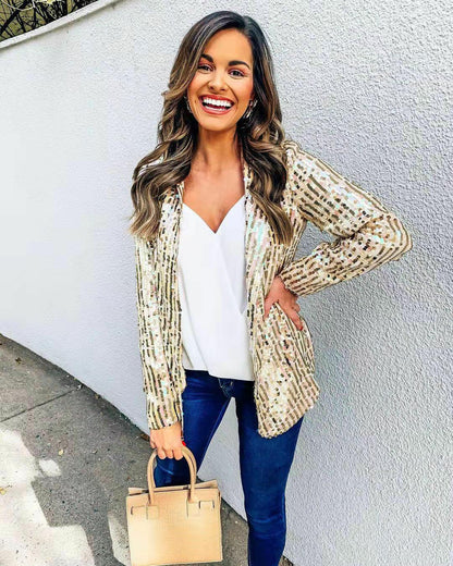 Frau in goldener Paillettenjacke, weißem Top, blauer Jeans, hält beige Handtasche, lächelt.