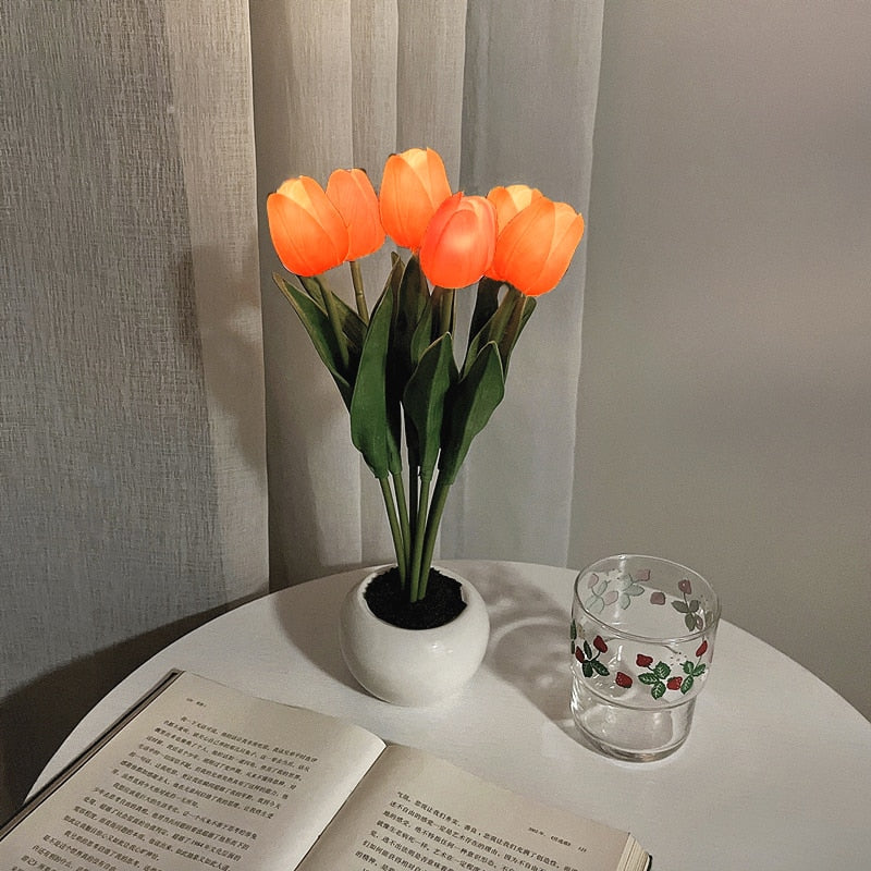 Künstliche orange Tulpen im weißen Topf auf Tisch, neben Buch und Glas, dekorative Beleuchtung.