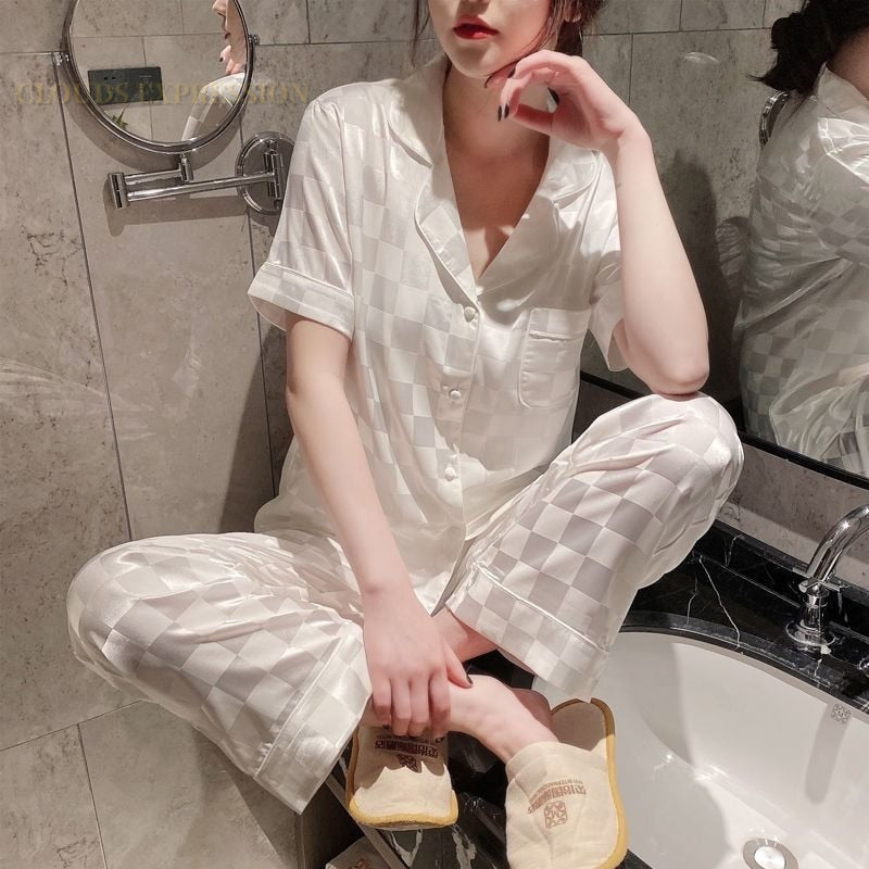 Frau in weißem Satin-Pyjama mit Karomuster im Badezimmer, sitzt auf Waschbeckenrand.