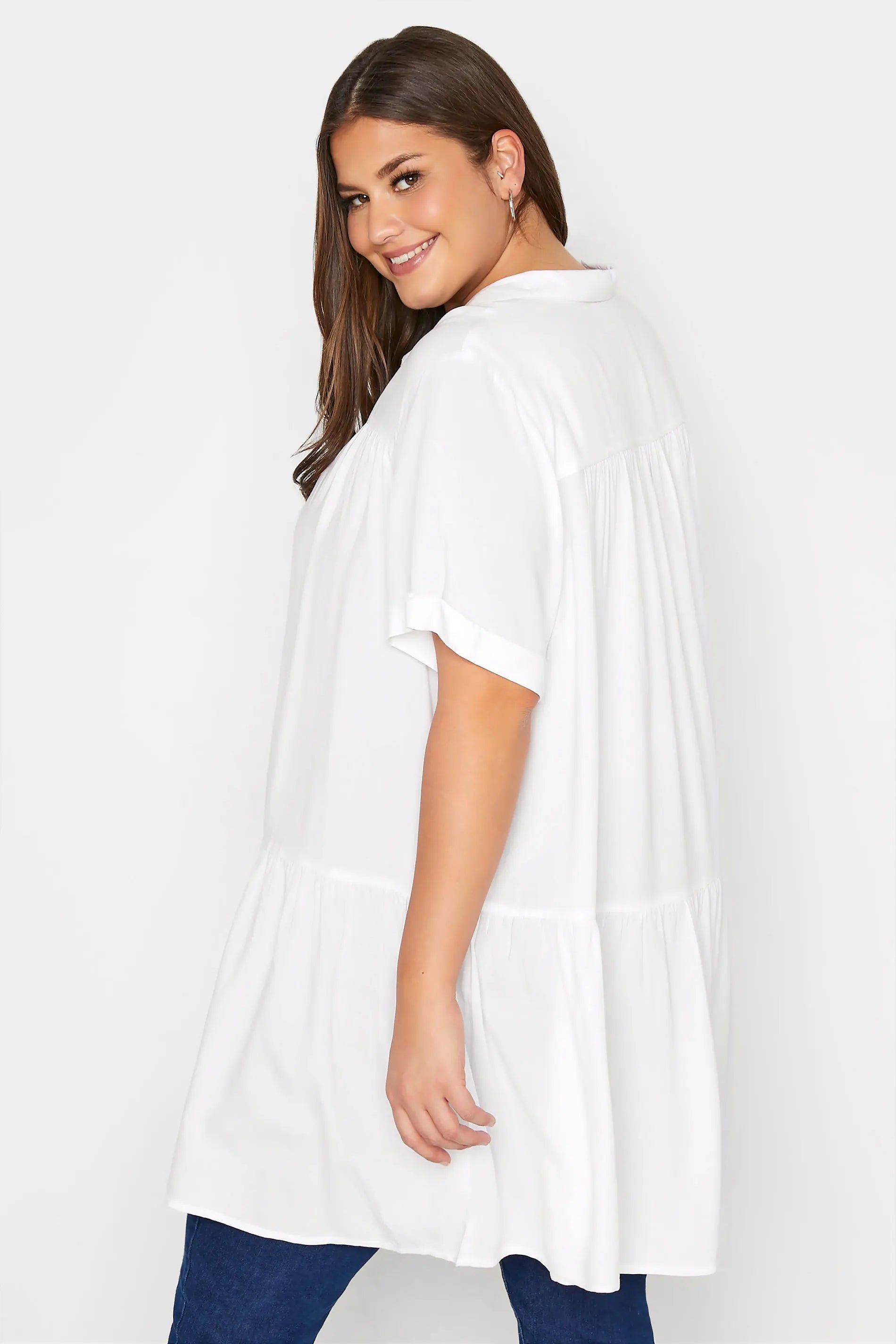 Frau in weißer Bluse, Plus-Size-Mode, lächelnd, seitlich, vor weißem Hintergrund.