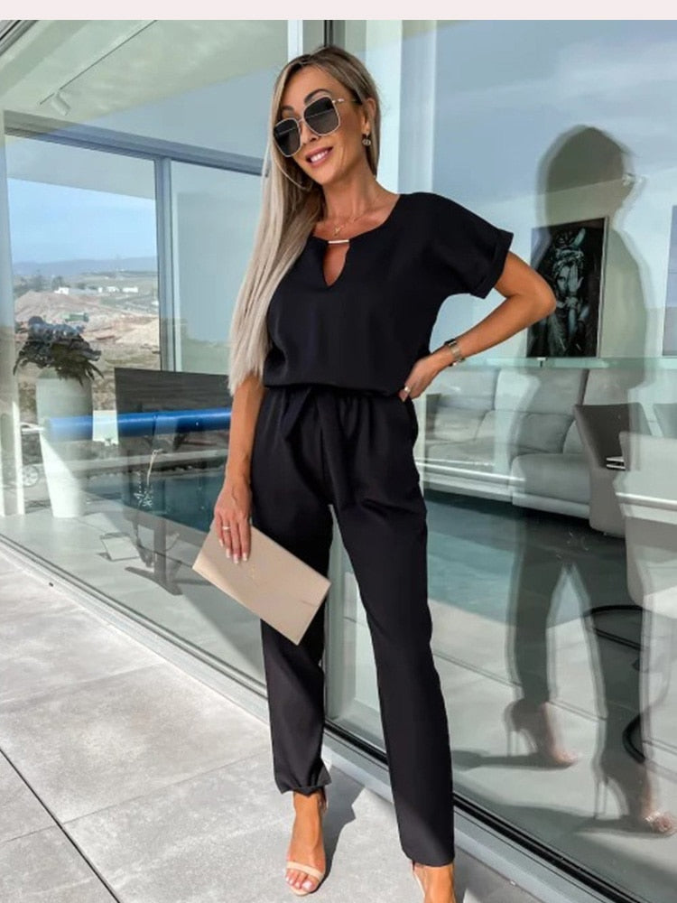 Frau in schwarzem Jumpsuit, Sonnenbrille, hält beige Clutch, moderne Terrasse im Hintergrund.