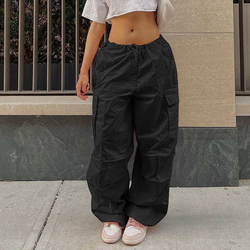 Frau in schwarzer Cargohose, weißem Crop-Top, rosa Sneakers, urbaner Hintergrund.