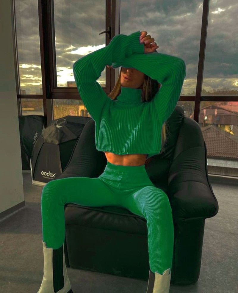 Frau in grünem Strickpullover und Hose, sitzt auf schwarzem Sessel, modernes Interieur.
