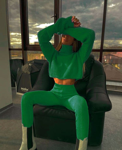 Frau in grünem Strickpullover und Hose, sitzt auf schwarzem Sessel, modernes Interieur.