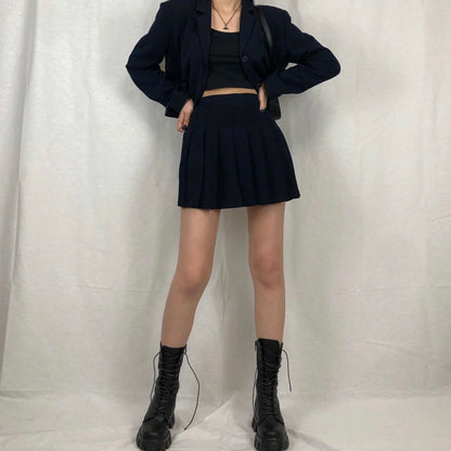 Frau in schwarzem Blazer und Faltenrock, schwarze Stiefel, modisches Outfit, weißer Hintergrund.