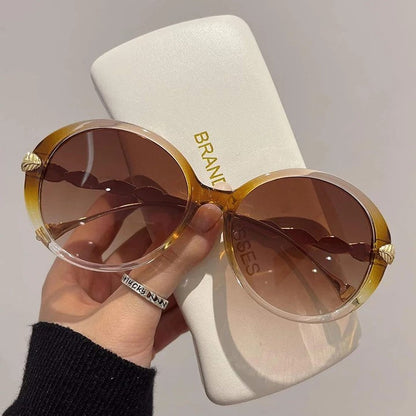 Große runde Sonnenbrille mit braunen Gläsern und goldenen Details, gehalten vor weißem Etui.