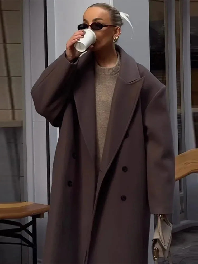 Frau in braunem Wollmantel trinkt Kaffee, trägt Sonnenbrille, modisch, urbaner Stil.
