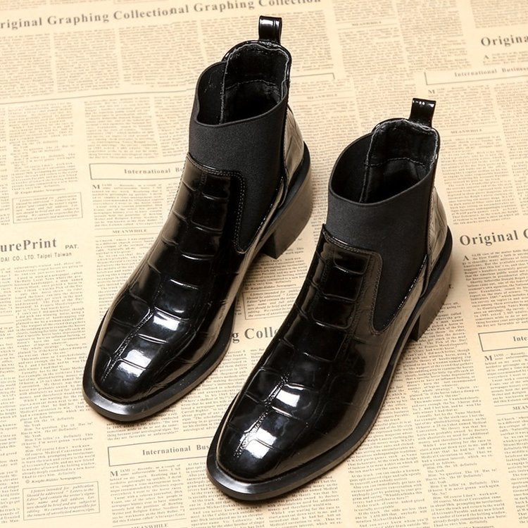 Schwarze Chelsea-Stiefel aus Leder mit Krokodilprägung auf Zeitungshintergrund.