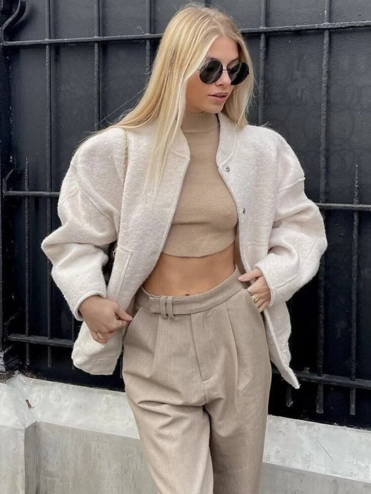 Blonde Frau in beiger Jacke und Hose, trägt Sonnenbrille, modisches Herbstoutfit, urbaner Hintergrund.