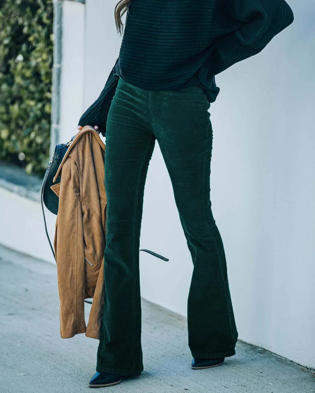 Frau in grüner Cord-Schlaghose, schwarzem Pullover, hält braunen Mantel, modisch, Herbstmode.