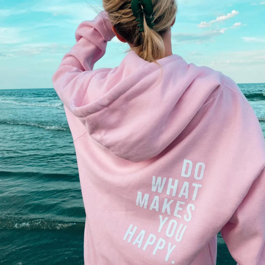 Frau am Strand in rosa Hoodie mit Aufschrift "Do What Makes You Happy", Meer im Hintergrund.