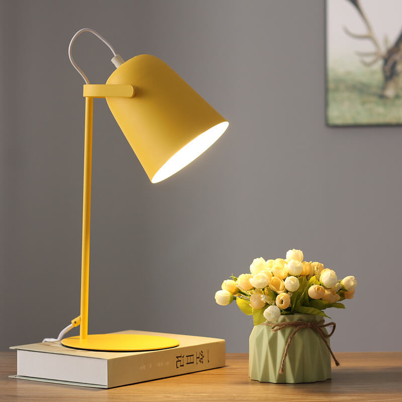 Gul bordslampa tänd på en bok vid väggen, bredvid en grön kruka med blommor och en tavla. 7