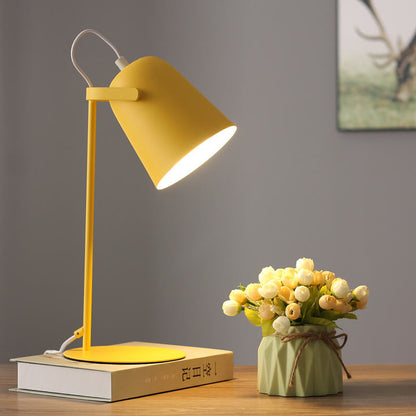 Gul bordslampa tänd på en bok vid väggen, bredvid en grön kruka med blommor och en tavla. 7