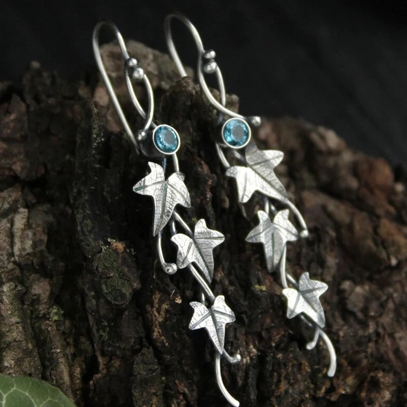 Silberne Ohrringe mit Efeublatt-Design und blauen Edelsteinen auf Holzuntergrund.