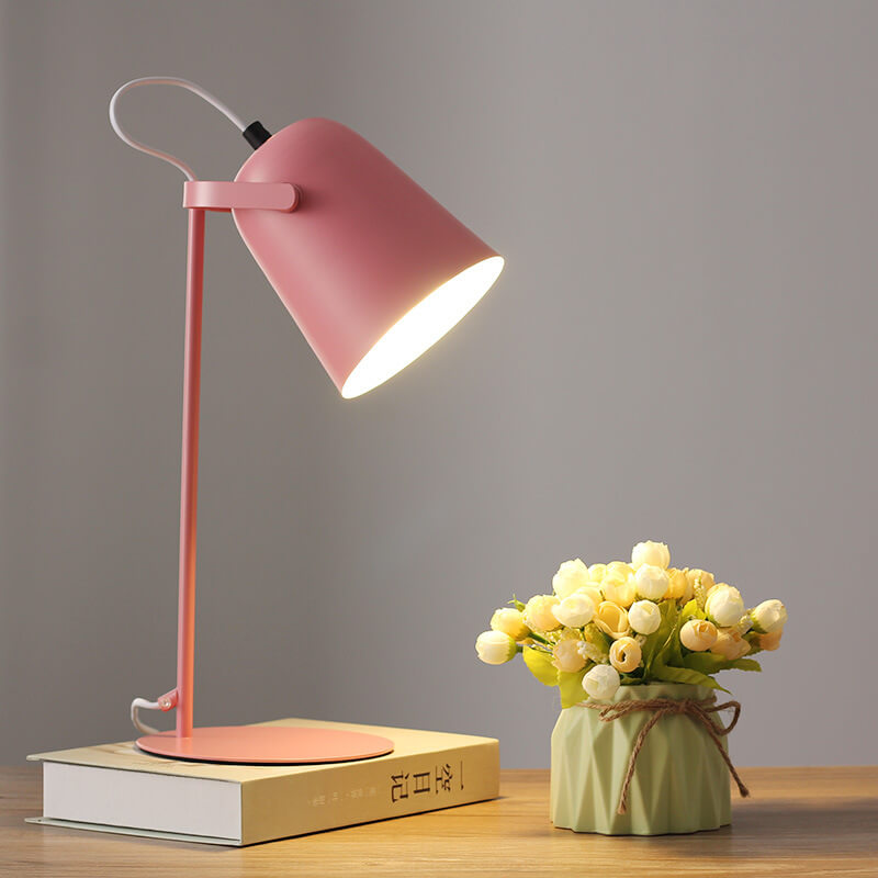 Rosa bordslampa med tänd skärm, placerad på en bok bredvid en grön kruka med vita och gula blommor. 8